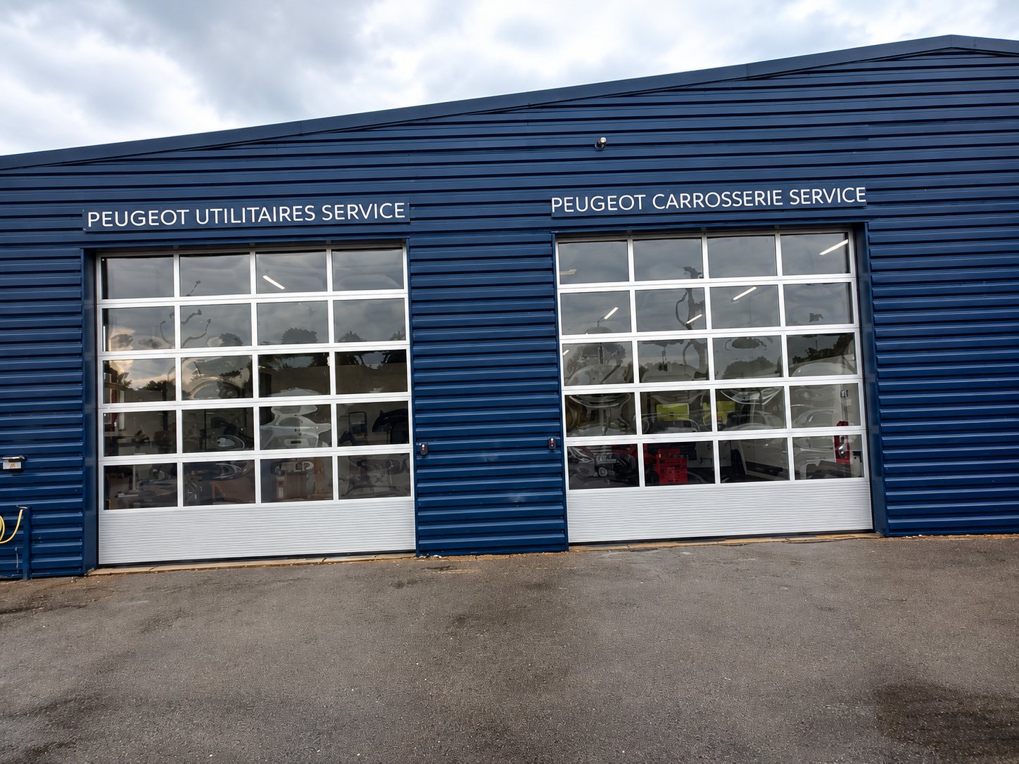 Façade d'un bâtiment bleu foncé abritant les ateliers Peugeot Utilitaires Service et Peugeot Carrosserie Service, avec deux grandes portes sectionnelles vitrées.