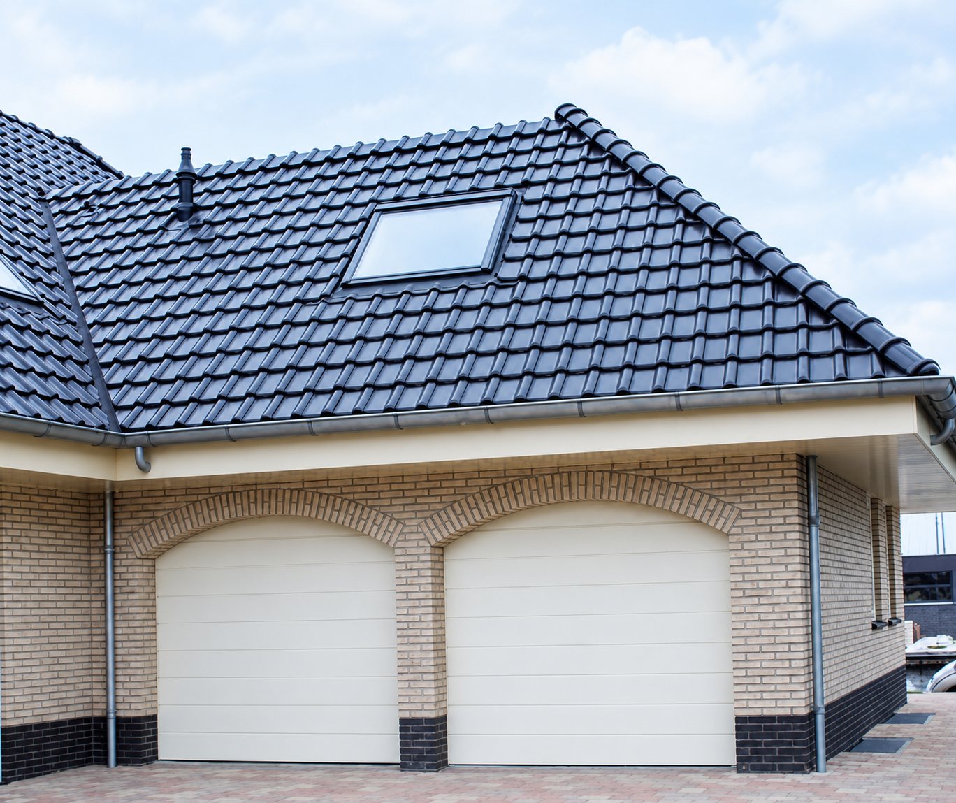 Un garage en briques claires avec deux portes sectionnelles crème à arc surbaissé, surmonté d'un toit de tuiles gris foncé avec une fenêtre de toit.