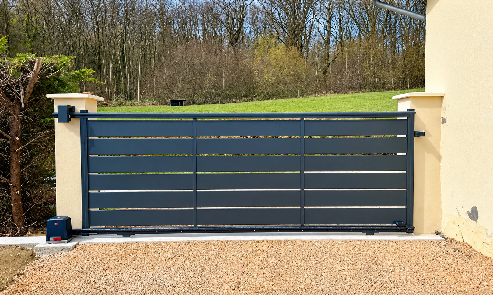 Un portail coulissant motorisé gris anthracite à lames horizontales, installé entre deux piliers beiges au bord d'une allée de gravier, avec une prairie et des arbres en arrière-plan.