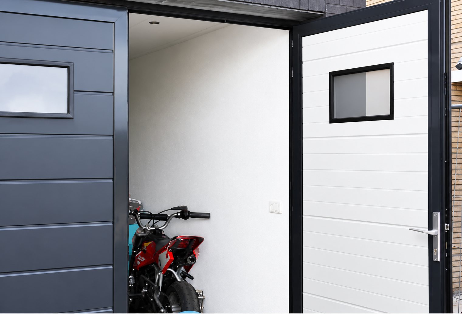 Une porte de garage sectionnelle anthracite à côté d'une porte blanche à hublot ouverte, laissant voir une moto rouge dans un garage aux murs blancs.