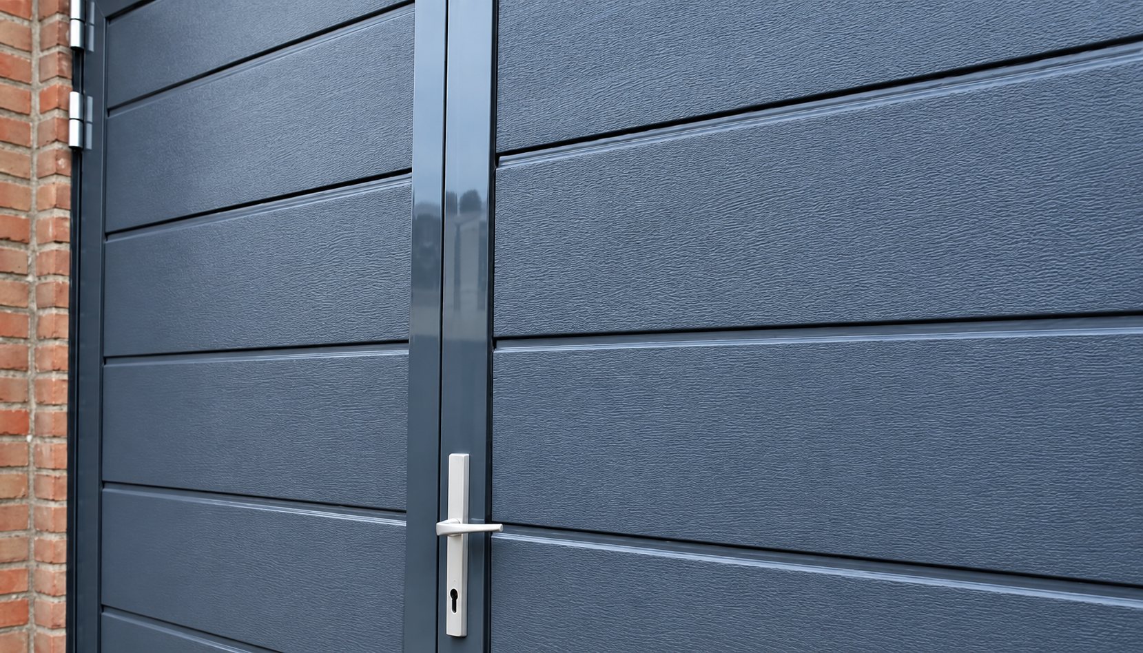 Gros plan sur une porte de garage battante gris anthracite à deux vantaux, avec panneaux horizontaux, poignée métallique et mur de briques à gauche.