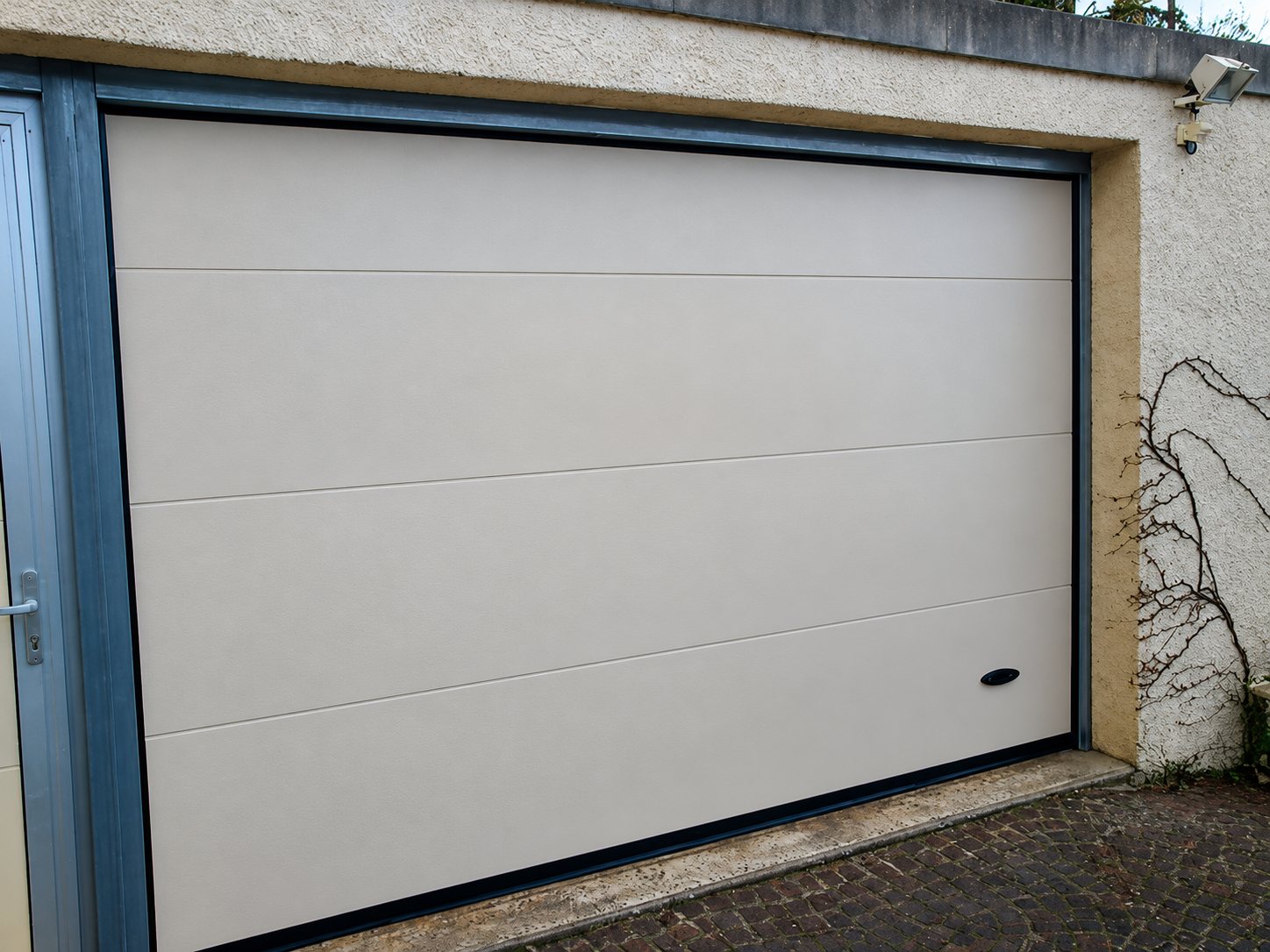Une porte de garage sectionnelle beige à panneaux horizontaux, posée dans un mur crépi avec un encadrement en pierre, devant un sol pavé.