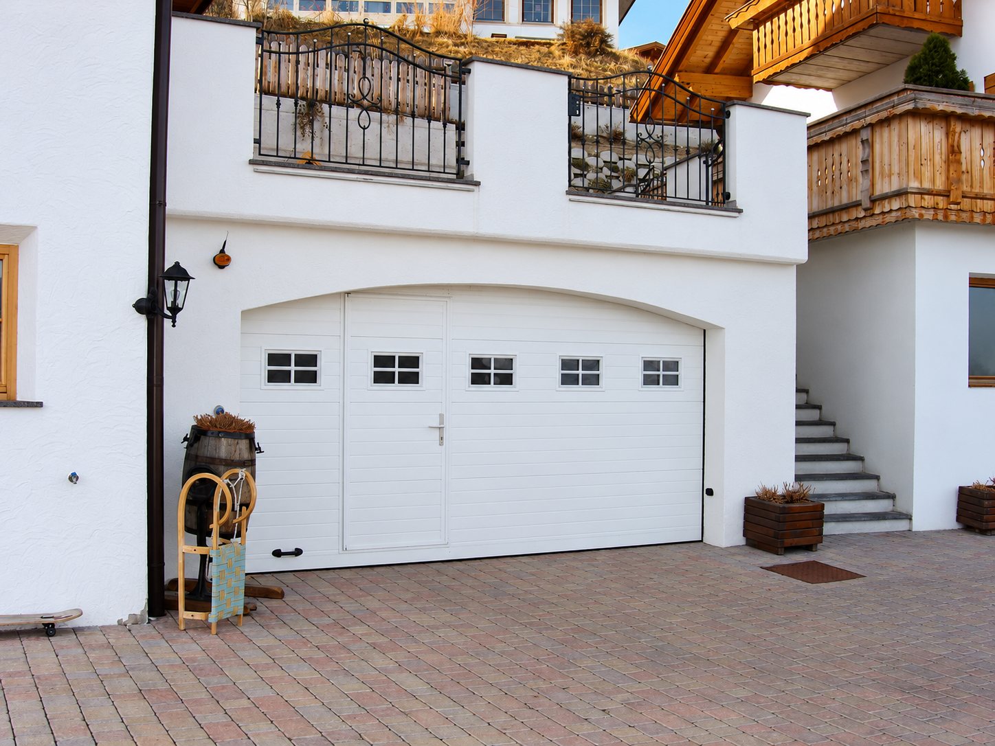 Une porte de garage sectionnelle blanche à linteau cintré, équipée d'un portillon et de petites fenêtres, sous un balcon en fer forgé, devant un sol en pavés.