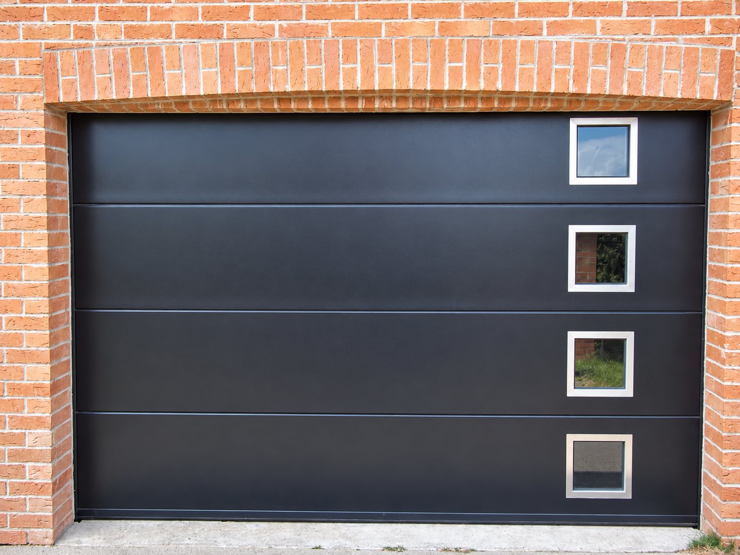 Porte de garage sectionnelle noire à quatre panneaux avec quatre petits hublots carrés alignés à droite, encadrée par un mur de briques orangées.