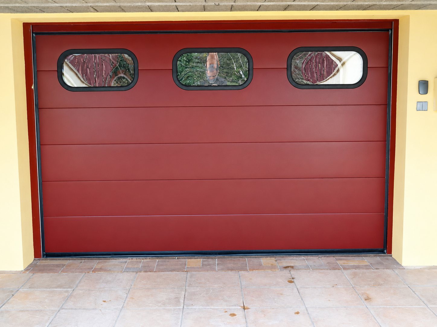 Porte de garage sectionnelle de couleur rouge bordeaux équipée de trois petits hublots, installée dans une façade jaune pâle.