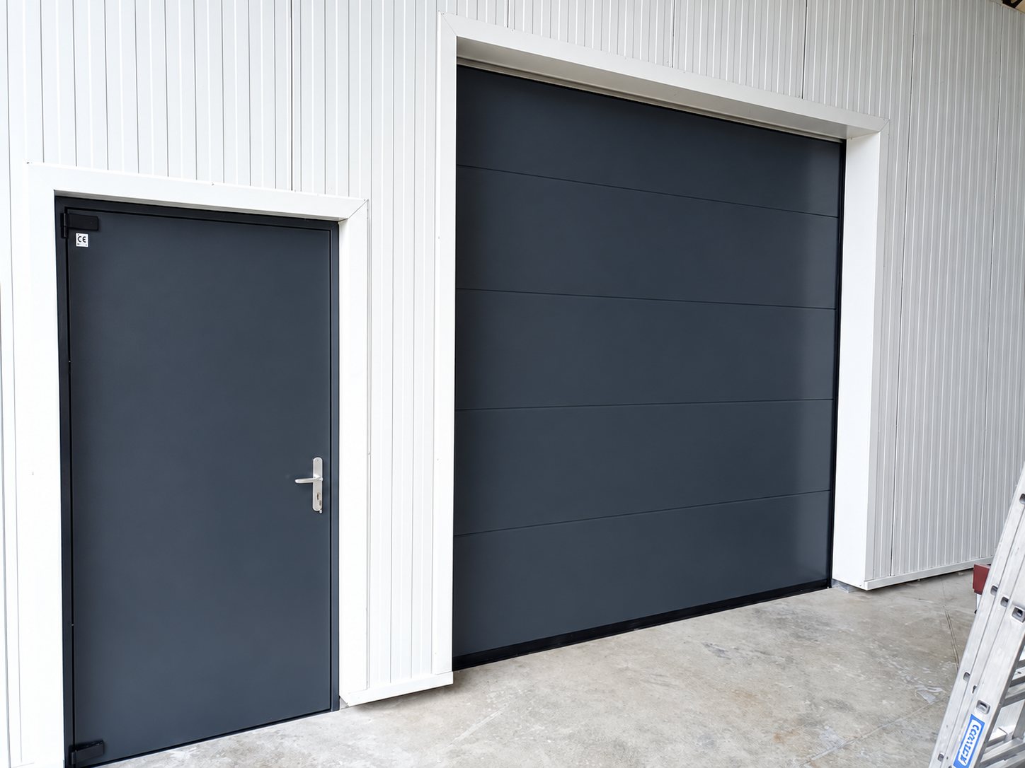 Une porte de service et une grande porte sectionnelle gris anthracite posées dans un mur de bardage blanc, avec une échelle en aluminium à droite.