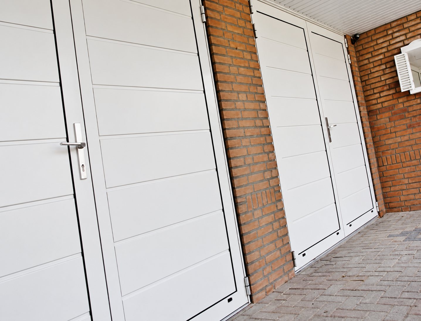 Plusieurs portes de garage battantes blanches à lames horizontales, encadrées de murs en briques rouges, sur un sol en pavés.