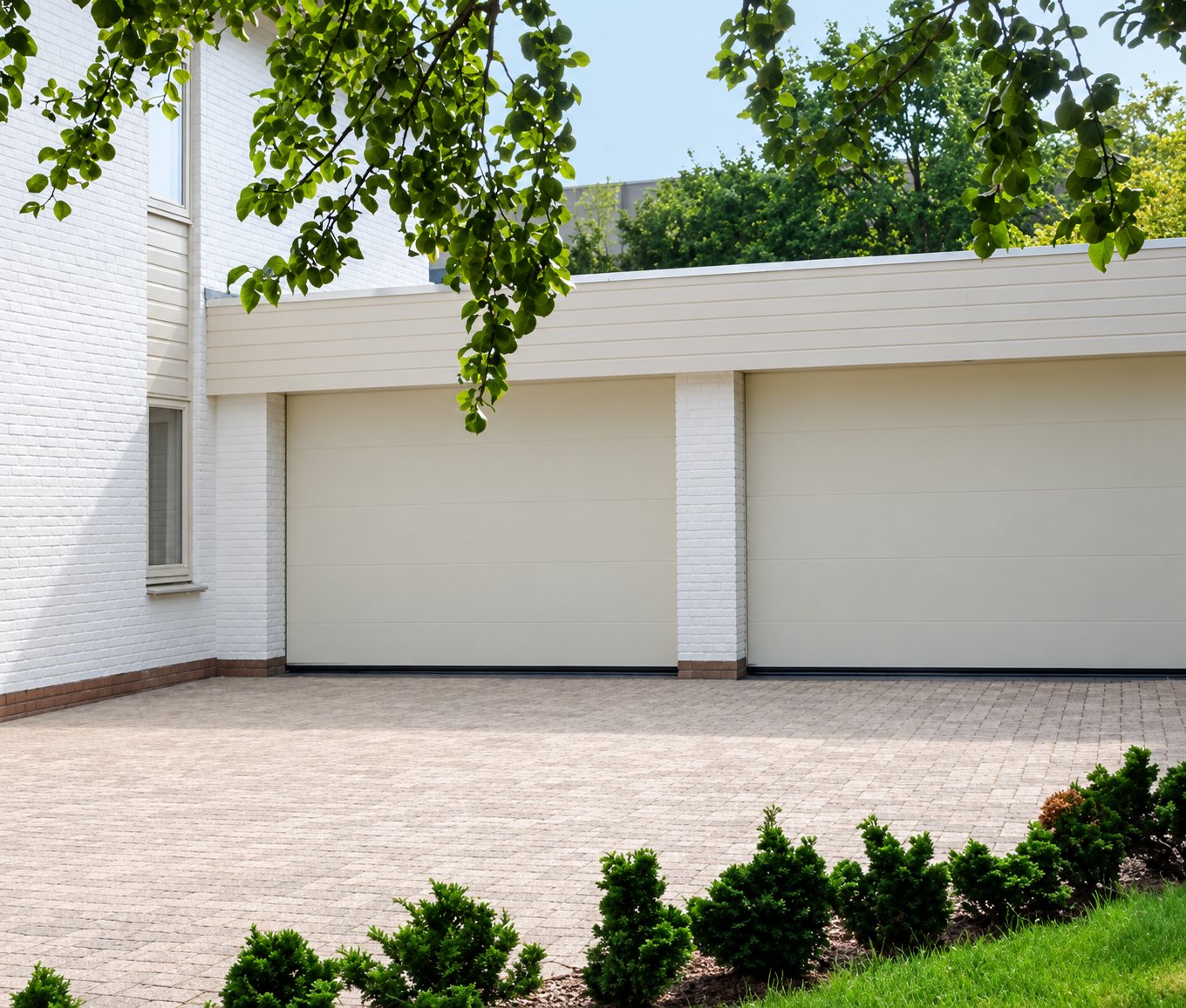 Deux portes de garage sectionnelles de couleur crème installées sur une maison blanche, devant une allée en pavés et des buissons taillés.